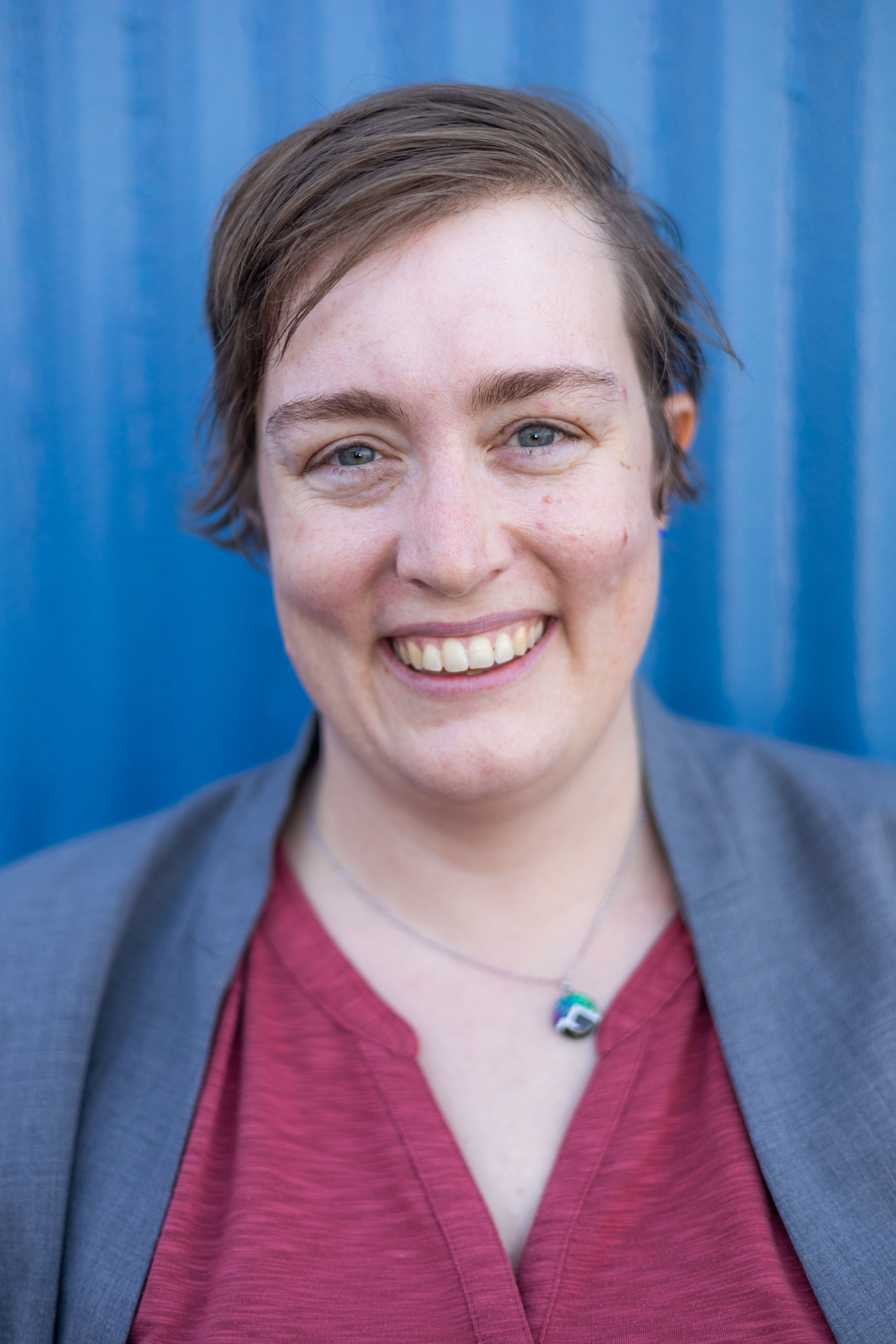 A white woman with short brown hair looks into the camera. She has gray eyes and a large smile showing her top row of teeth. She's wearing a gray colorless blazer and a red shirt with a split v neck. Her round pendant necklace shows two mountains with the aurora borealis above them. The background is royal blue.