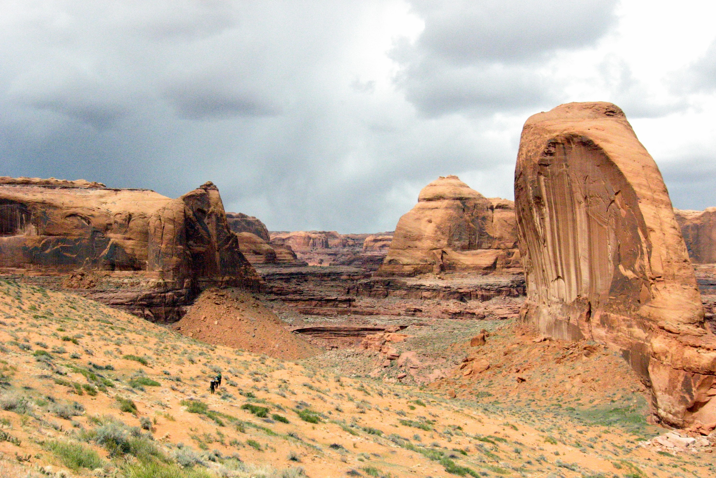 The foreground is yellow-ish sand with small, occasional tufts of green grass. Ther eare three tiny hikers in that area in the distance. The middle of the photo has giant rock structures, carved by millions of years of water. You can clearly see the layers in the distant rocks which are cliff like. There is an avalanche of stone beyond the hikers. On the right side is a giant stone that looks like what a gravestone would look like to an ant. It is rounded on top, and less on the side.s There are a number of vertical sediment stripes in that rock. The sky is cloudy and dark to light gray.