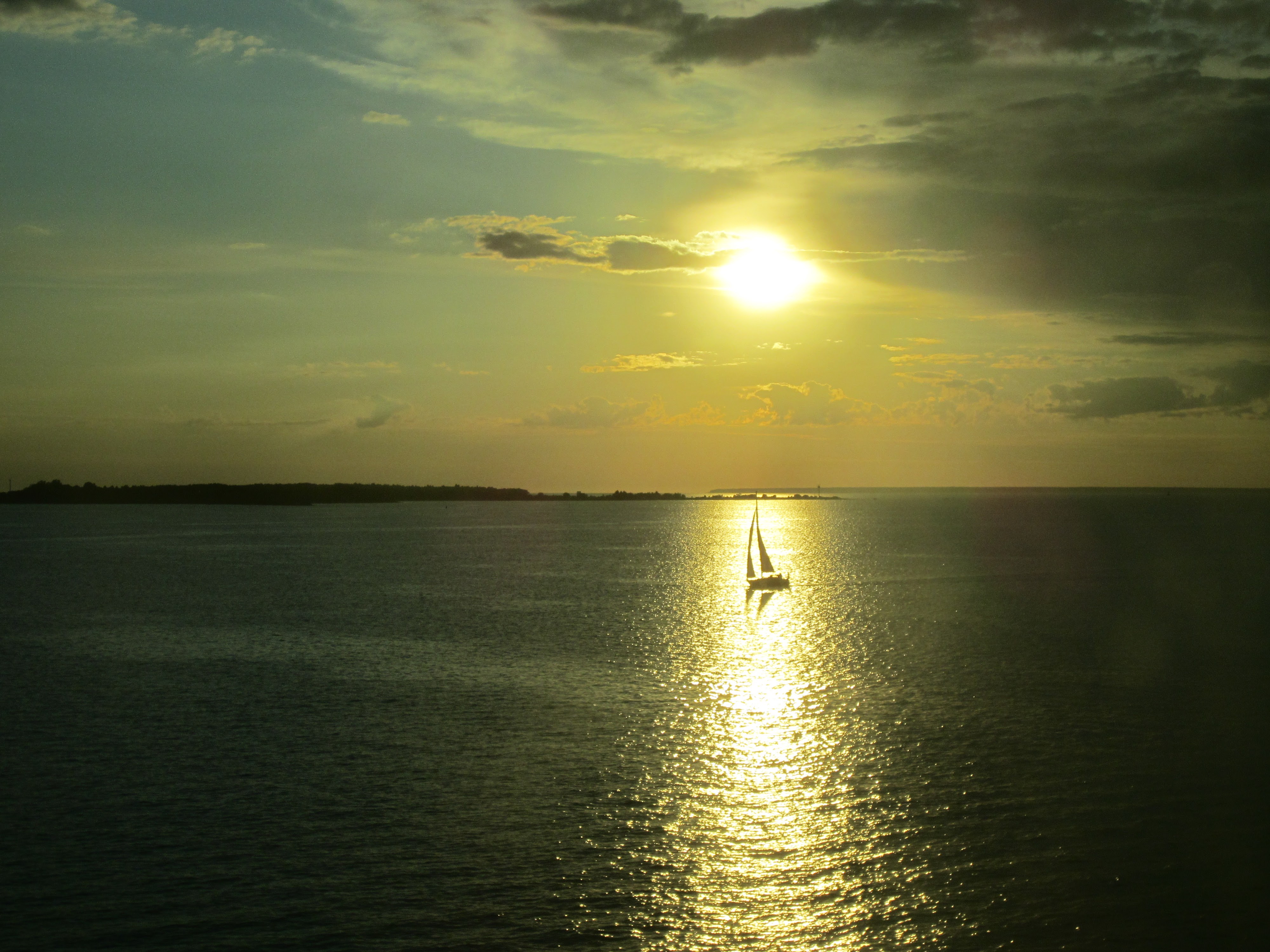 The picture was taken at dusk. Two thirds of the way to the right in the bottom is a sailboat. No details of the sailboat can be seen because the sailboat is in the path of the sun, which is directly above the boat in the photograph. The sun is reflecting off the dark ocean with the sailboat in the direct path. There are several dark clouds off to the right. On the left, a point of land sticks out just touching the top mast of the small boat.