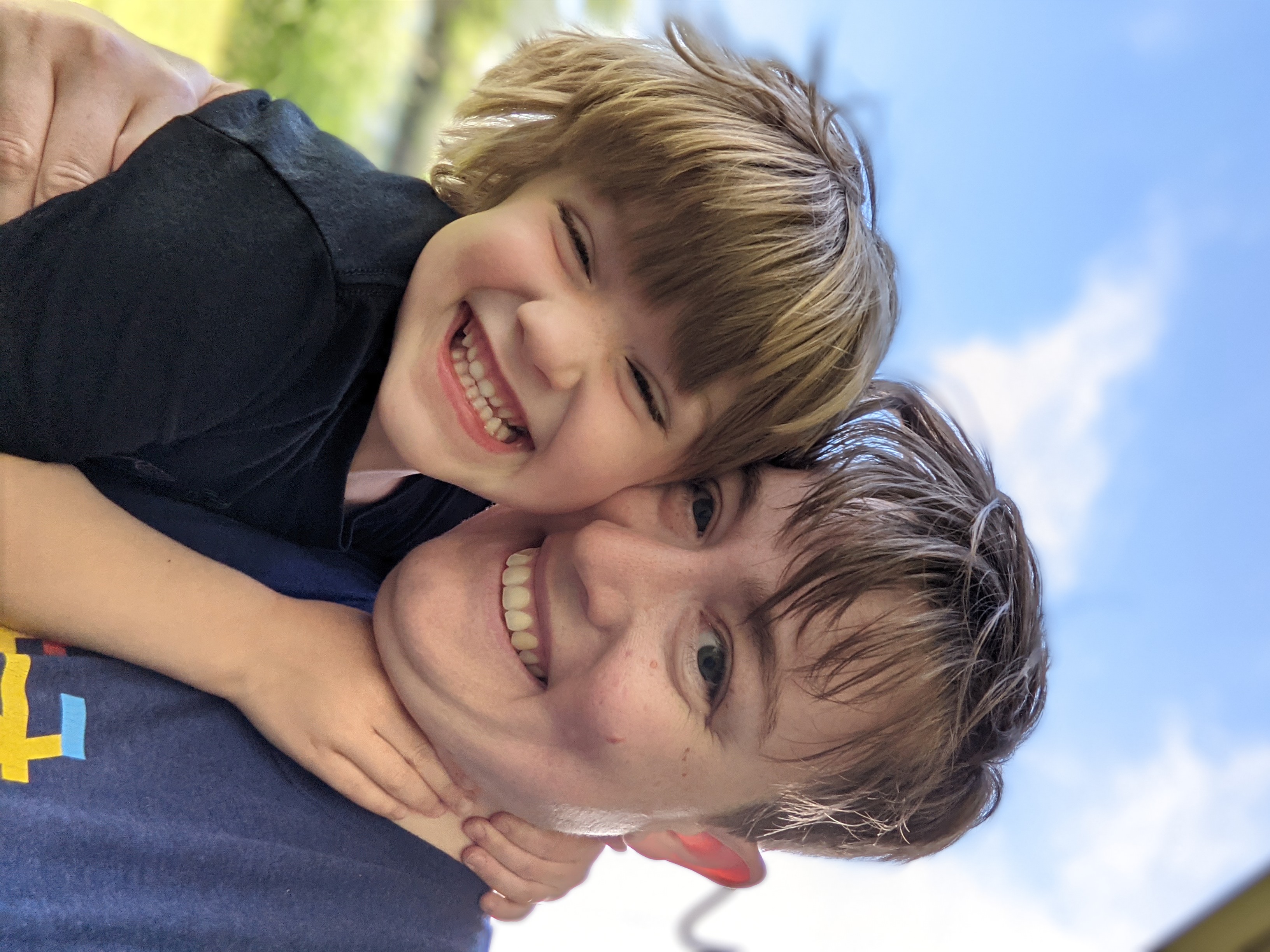 A white middle-aged woman, Katie, is on the right. She has messy short brown hair and a big smile. She is wearing a blue tshirt. On the left is her 5 year old daughter Lily wearing a black t shirt, a giant grint, and messy blonde short hair. Her hands are grasping Katie's neck.